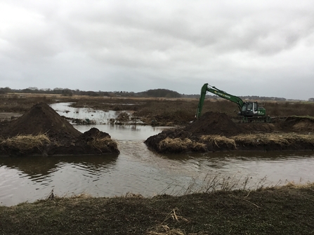 The breach in the River Alt embankment with Lunt Meadows in the background. An amphibian digger (pictured) has been used to ready the breach for the sandbags to be placed, and level out the gap. Water is slowly pouring out of Lunt Meadows and back into the River Alt