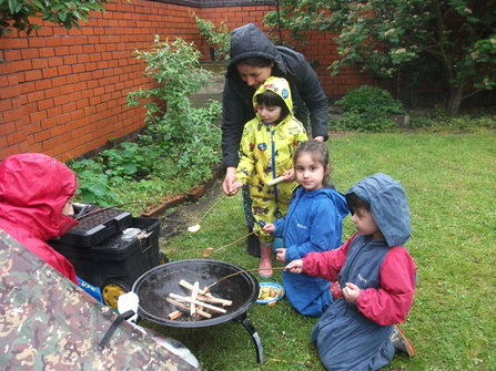 Three children in waterproofs cooking over a fire with an adult on a rainy day