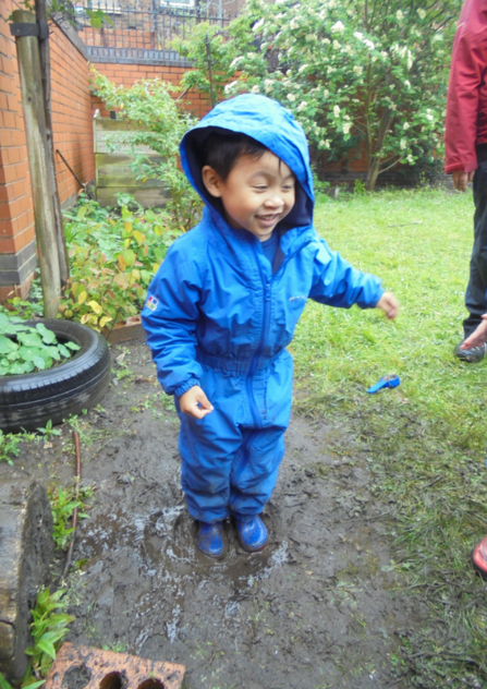 A child wearing a blue waterproof all-in-one suit, splashing in a muddy puddle and smiling on a rainy day