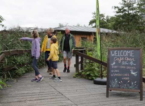 An older couple and two children walk on the bridge at Mere Sands Wood. A welcome sign sits nearby.