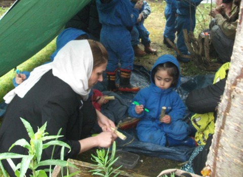 Children in blue all-in-one suits and adults working together on natural crafts under a tarpaulin