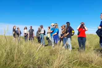 A group of people standing in a line across a grassy sand dune and looking into the distance. The sky is bright blue