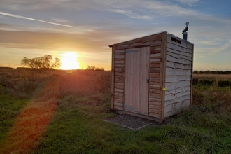 A wooden shed-like building housing a compost toilet standing amongst vegetation at Lunt Meadows as the sun sets behind it