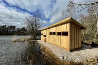 A wooden bird hide standing on a pebbly shore next to a lake at Mere Sands Wood
