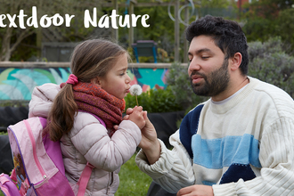 A father and daughter blowing on a dandelion clock against a park backdrop