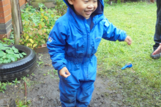 A child wearing a blue waterproof all-in-one suit, splashing in a muddy puddle and smiling on a rainy day