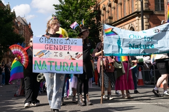 A lgbt+ pride parade with a banner saying 'out for nature' and a trans flag saying 'changing gender is natural'