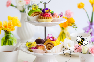 A cup of tea, colourful cakes on a cake stand with yellow, white and pink flowers in vases. A cartoon bee is flying past.