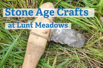 A middle stone age hand axe made from flint rock with a wooden handle and nettle string lies in the grass. Blue text over the top of the image says stone age crafts at lunt meadows