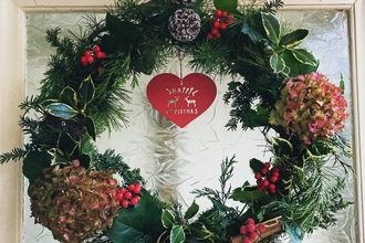 A wreath on a glass door, it is green and made with holly, pine cones, hydrangea, red berries and a decoration in the middle that says merry Christmas