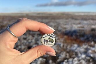 A sunny moorland with a hand holding a bog squad badge