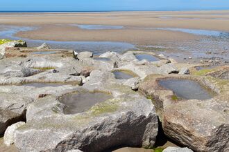 Pools of water are seen in rocks which form part of the rocky groyne sea defence. The tide is out and sand can be seen behind the rocks