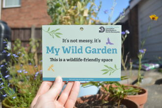A My Wild Garden plaque being held up in a garden with pots of flowers visible in the background
