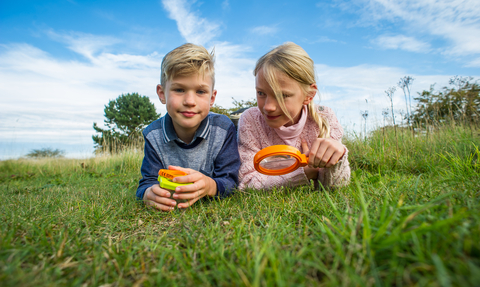 Children laying on grass with magnifying glass with blue sky