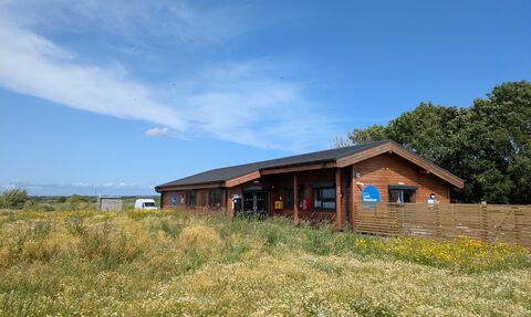 The Learning Centre, a wooden single storey building with a wildflower meadow in front and a fence. There is a blue sky above.