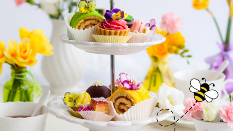 A cup of tea, colourful cakes on a cake stand with yellow, white and pink flowers in vases. A cartoon bee is flying past.