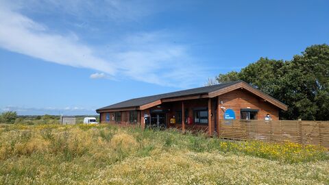 The Learning Centre, a wooden single storey building with a wildflower meadow in front and a fence. There is a blue sky above.