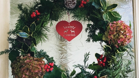 A wreath on a glass door, it is green and made with holly, pine cones, hydrangea, red berries and a decoration in the middle that says merry Christmas