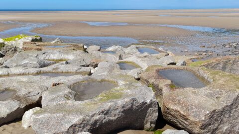 Pools of water are seen in rocks which form part of the rocky groyne sea defence. The tide is out and sand can be seen behind the rocks