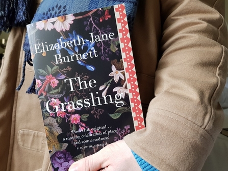Someone wearing a coat and scarf holding a copy of The Grassling