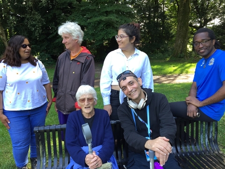 A group of Myplace participants standing around a bench at Witton Park, talking to each other and smiling at the camera