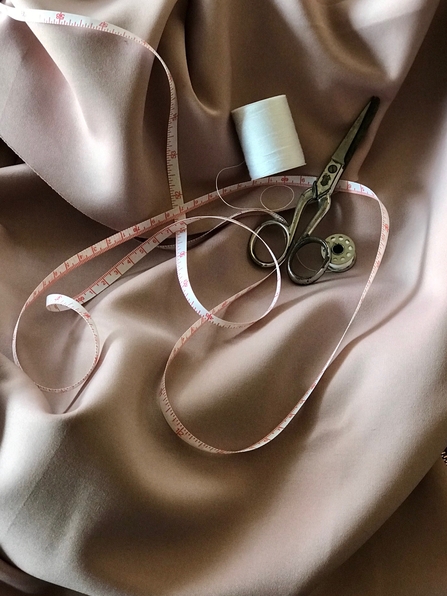 A pair of scissors, a spool, sewing thread and a tape measure on top of pale pink silk