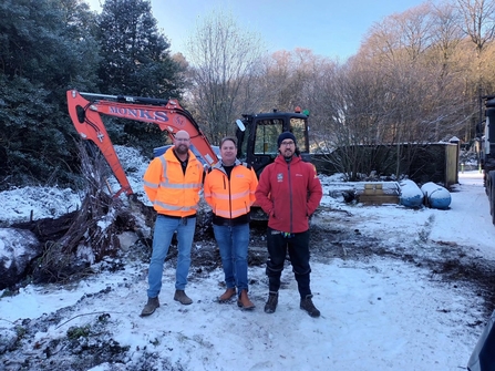 Three people in the snow standing in front of a digger