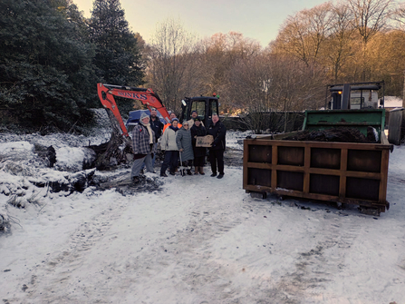 A group of 8 people stood in the snow in front of a digger