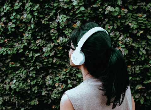 A woman wearing headphones and standing in front of a hedge outside