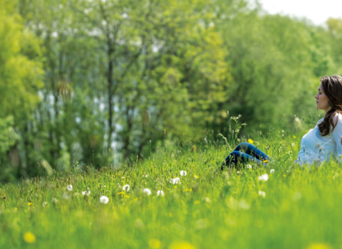 A woman sits in the grass with her eyes closed, surrounded by trees