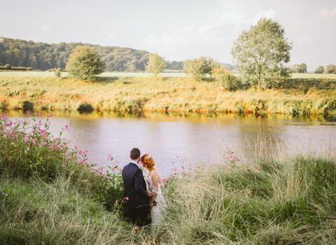 A bride and groom stand on the riverbank looking out over the river Ribble