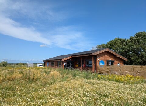 The Learning Centre, a wooden single storey building with a wildflower meadow in front and a fence. There is a blue sky above.