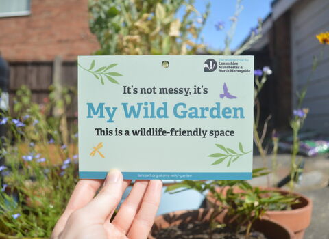 A My Wild Garden plaque being held up in a garden with pots of flowers visible in the background